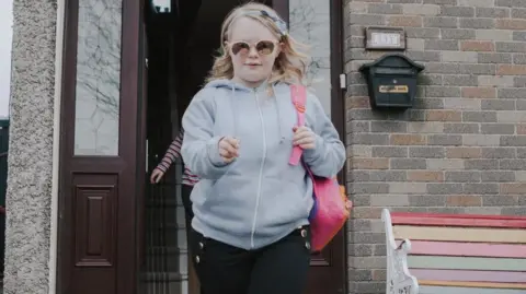 Claudia walking towards the camera wearing a pair of black jeans, a grey zip-up hoodie and white trainers. She has blonde hair, pink sunglasses and a pink backpack. In the background is a house with the front door open.