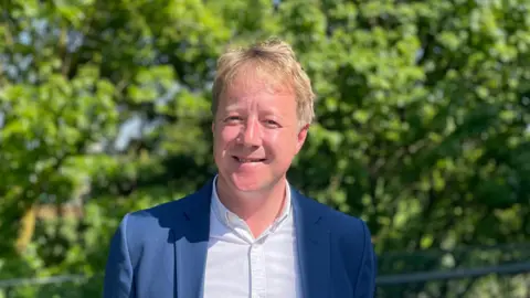 Kate Moser Andon/BBC Paul Bristow wearing a blue suit and light blue shirt, with foliage in the background