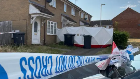 Blue and white police tape cordoning off the entrance to the house, which is mid-terrace and made of cream bricks. There is a white tent surrounding the entrance.