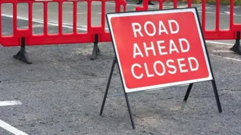 A red road sign reading "Road ahead closed" in the middle of a streey. There are red street barriers behind it.