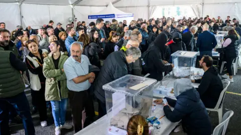 Getty Images People wait in several long lines in a large white tent to cast a vote in plastic ballot boxes.