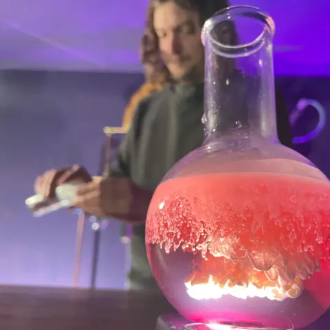 A glass flask sits on top of a light, illuminating three different liquids that are coloured purple, pink and white which are not mixing. In the background Roy prepares the next experimental drink.