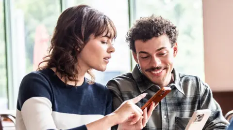 A woman with dark shoulder-length hair points at a smartphone held by a smiling man with curly hair and a moustache.