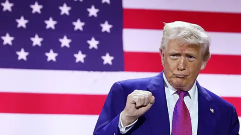 US President Donald Trump pumps his fists on stage with an American flag seen behind him as he tours the Al Udeid Air Base on 15 May 2025, in Doha, Qatar.
