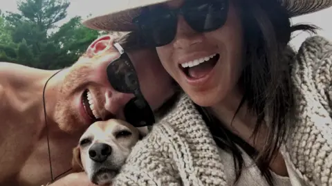 Prince Harry and Meghan, The Duke and Duchess of Sussex A handout photo shows the Duke and Duchess of Sussex with their late dog, Guy. The two are seen leaning together as the dog pokes his head between their shoulders and looks towards the camera. 