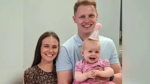 A family photo of Harry Foster, his wife and daughter Olivia. All are smiling and baby Olivia is wearing a little party hat with a pink pom pom on. 