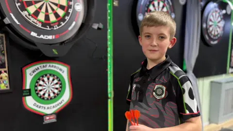 BBC Jac, 12, stands next to a dartboard at Cwm Taf Arrows Darts Academy and holds his darts