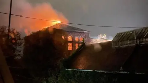 Roxy Wilkes Orange flames can be seen through the pumping station's high arched windows and fire is coming out through the building's pitched roof. It is night time. 