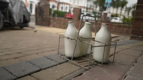 Three glass milk bottles stand in a metal holder on the doorstep of a house. Beyond them is a driveway leading to a gate. And in the background are residential properties. 