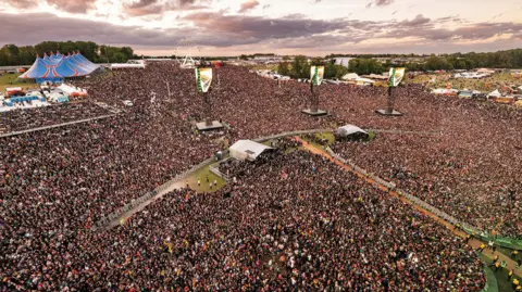 Sarah Louise Bennett/Download Festival An aerial shot of thousands of people at a festival, with a large blue performance tent in the top left and tall sound speakers in the crowd.