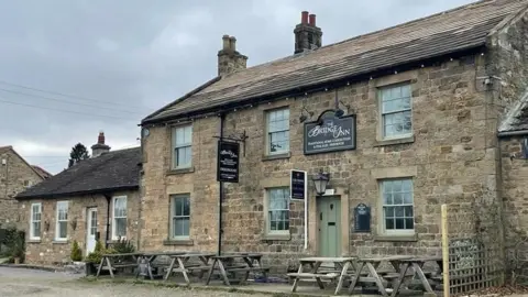 LDRS A general view of The Bridge Inn in Whorlton. It is a yellow sandstone two-storey building with traditional sash windows and a mint green wooden front door. There is a smaller one storey cottage-type building to one side and there are picnic tables outside the pub.