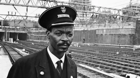 A black man in a smart uniform and hat is pictured in front of railway lines.