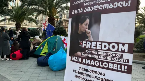 A poster with a picture of Mzia Amaglobeli sites alongside a tent camp in Batumi