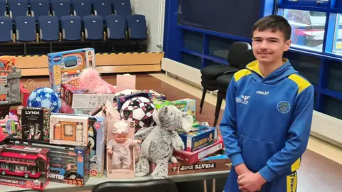 Luke, dressed in a Newcastle Eagles basketball tracksuit, stands next to a table full of toys he has collected for children in care.