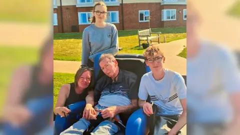Supplied A family photo with a man in a wheelchair in the centre, with a young boy to his right, wife to his left, and daughter stood behind.