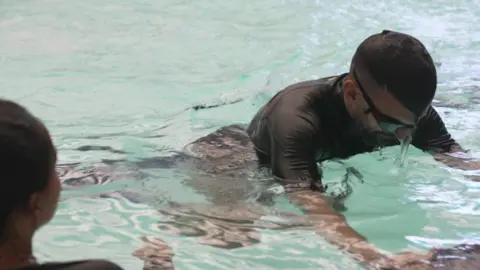 A man is putting his face into the water at a swimming pool. He is wearing goggles.