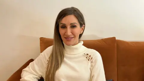 A woman with long dark hair wearing a white knitted jumper sits on an orange sofa. She is looking directly at the camera. 