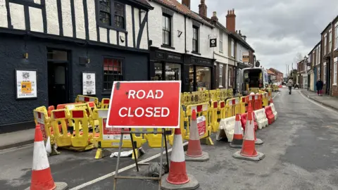 A red road closure sign stands in front of a tangle of yellow barriers on a narrow road. A medieval pub and a row of shops line the street.