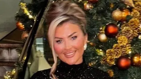 Taylor Jenkins is standing in front of a Christmas tree and smiling brightly at the camera. Her blonde hair is pulled into a bun and she is wearing a sparkly black dress and earrings.