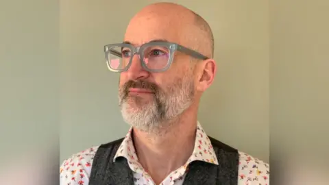 Cheltenham Science Festival Mark Miodownik MBE. He has grey facial hair and reading glasses with grey frames. He is looking to the side of the frame.
