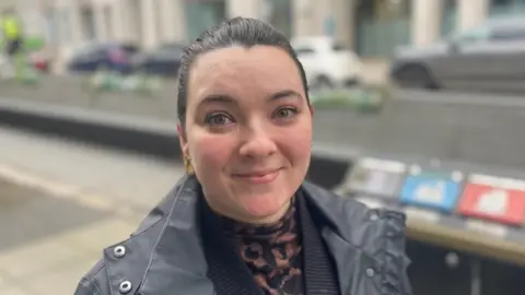 Sophie Crawford is smiling. Her dark hair is tied back, She's wearing a black coat, a black cardigan and a tortoiseshell top. She's in a street. The background is blurred. Some bins and cars are behind her. 