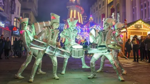 Handout Five drummers dressed in silver outfits, with their faces painted white. They are holding drums that are lit up. People are surrounding them in the street. Fairgrounds rides can be seen in the background. 