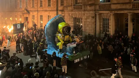 George Carden People stood sround an effigy of a yellow hazmat suit sat in a wave with the words SWIM IN POO on the front of the float. People are holding fire torches at the back of the effigy