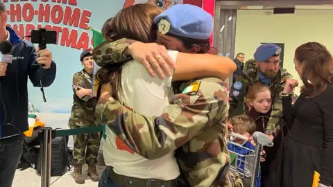 Two women hug in the airport. One woman is a solider in camouflage clothing with a blue hat. She is hugging a woman in a white t-shirt with an irish flag. In the back there are more soldiers with their families.