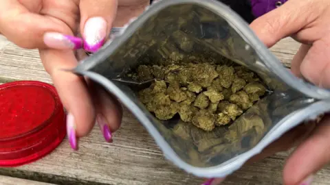 A silver pouch filled with Medical Cannabis prescription, dried flower. There is also a red grinder in the background. 