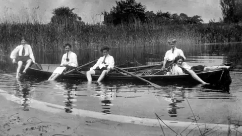 BPEOSA collection Five women, in Edwardian white shirts and skirts, with ties round their necks in a long rowing boat. Four women have their skirts rolled up with their feet in the water, while the fifth is sitting fully inside the boat. Reeds are at the bank behind them. 