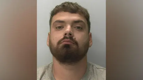 Devon and Cornwall Police Ben Helliker - aman with a dark beard, moustache and short dark hair is wearing a grey T-shirt. He is looking at the camera with slightly downcast eyes.