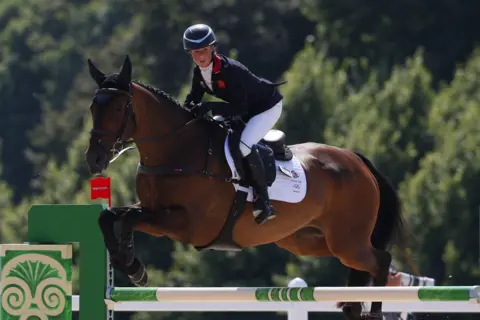 Getty Images A female rider on a brown horse clears a jump during a showjumping round. She is wearing a black jacket, white jodhpurs and a black riding helmet. The jump has green and white barriers. Green trees are in the background.