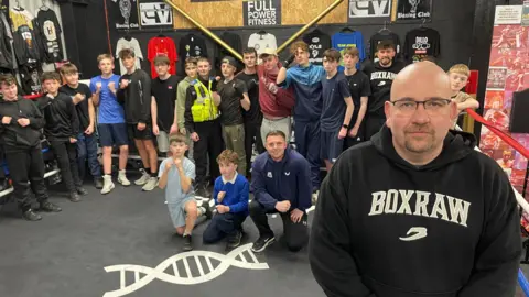 Fifteen young men, a police officer and four adults are stood in a boxing ring. One adult with a shaved head, glasses and black hoodie printed with the word 'BOXRAW' stands in front of the crowd. The boxing ring is black with a white image of a helix. There are posters and t-shirts pinned to the walls.