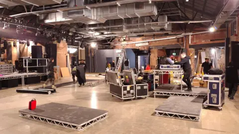 The inside of a music venue. Various flight cases are stacked around the room as people work to put the finishing touches to the building. 