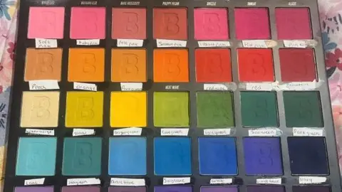 Ailish McBride There is a black make-up palette containing eyeshadow squares of different colours. Each row has different shades, including red, yellow, green, blue and pink. Underneath each square is a small white label that has the name of the colour. The palette is resting on a pink sheet with flowers on it.