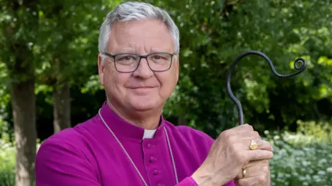 Stephen Lake, dressed in his deep purple robes, with a silver chain, gold rings and black-frames glasses, holds a black metal staff and smiles at the camera in a garden. 