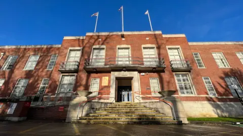 An exterior shot of a red-brick council building. The shot is taken from a low angle, and shows a staircase into the building, and flags at the top. There is a blue sky above.