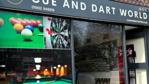 Jenny Phillips The outside of the shop, with large images of darts, snooker and pool. The name Cue and Dart World is at the top of the shop and the front door is open on the right.