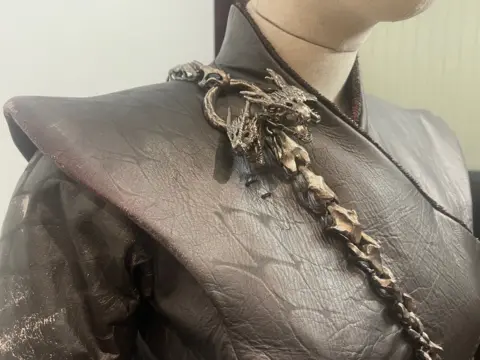 This close-up image shows a detailed leather Daenerys Targaryen costume. The dark, textured material is adorned with a metal chain featuring dragon designs.