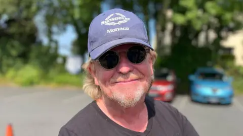 Deputy Bopb Murray wearing a blue hat and black sunglasses and smiling into the camera. He has long ginger hair and a ginger and grey beard.