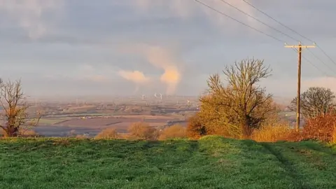 Rispo/BBC Weather Watchers A distant view of countryside from the Cotswolds in Gloucestershire, under winter sunshine. On the horizon is a distinct plume of white cloud rising up high into the sky