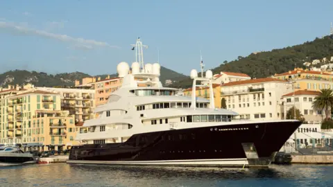 A large burgundy and white coloured yacht is moored in a harbour in the south of France with smaller vessels alongside it. A number of white, yellow and cream-coloured buildings are visible in the background and are set against a blue sky.