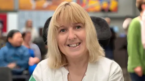 A woman, probably in her early 40s, smiles. She has a blonde, slightly wavy bob, and is wearing a white tunic style top. There are blurred out people sat behind her.