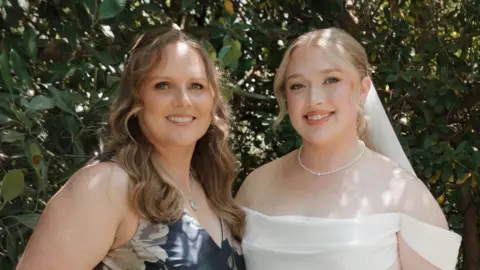 Hannah and her mother at Hannah's wedding. Her mother is wearing a blue dress with white flowers. Hannah on the right is dressed in a white wedding dress and veil