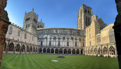 A panorama shot of a green in the centre of Durham Cloisters, with three towers visible beyond it