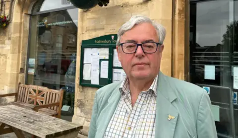 A man in a white shirt and green jacket stood in front of a building with an information board behind it.