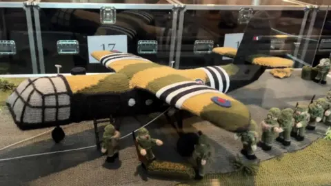 A green knitted plane with knitted soldiers lined up next to it.