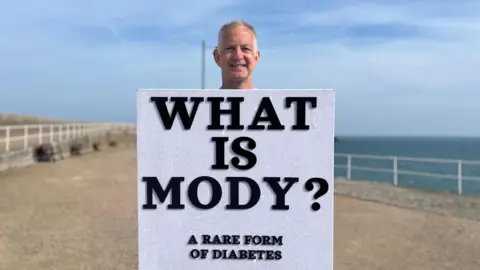 Richard is standing on a seaside promenade wearing a sugar cube that reads ‘WHAT IS MODY? A RARE FORM OF DIABETES,’ with the sea and railings in the background under a blue sky