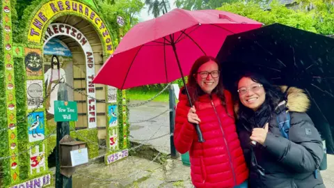 Two women hold umbrellas beside a natural water spring. Above it is a large board depicting Jesus. The design is bright and colourful, made from flower petals and natural materials. The words "The resurrection. I am the resurrection and the life. John 11 V25-26" are also included in the design.