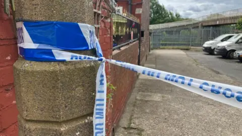 BBC/Phil Bodmer Police tape tied around a lamppost, with a bridge in the background.